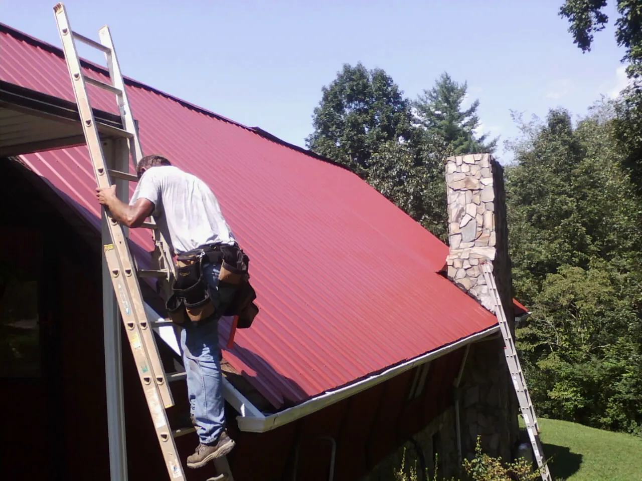 Roofer on a ladder beside a red metal roof and stone chimney — Soffit & Fascia Repair by Roofing Brothers in North Haven